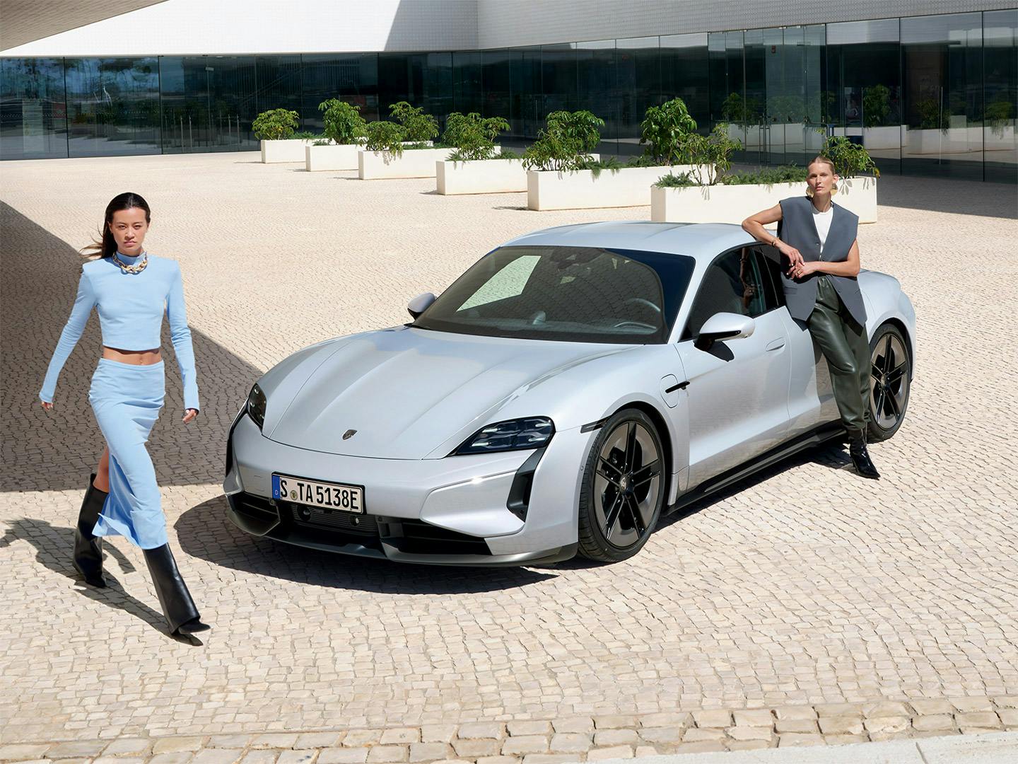 Eine Frau, die an einem Porsche Taycan lehnt. Eine weitere Frau läuft vorn am Fahrzeug vorbei.