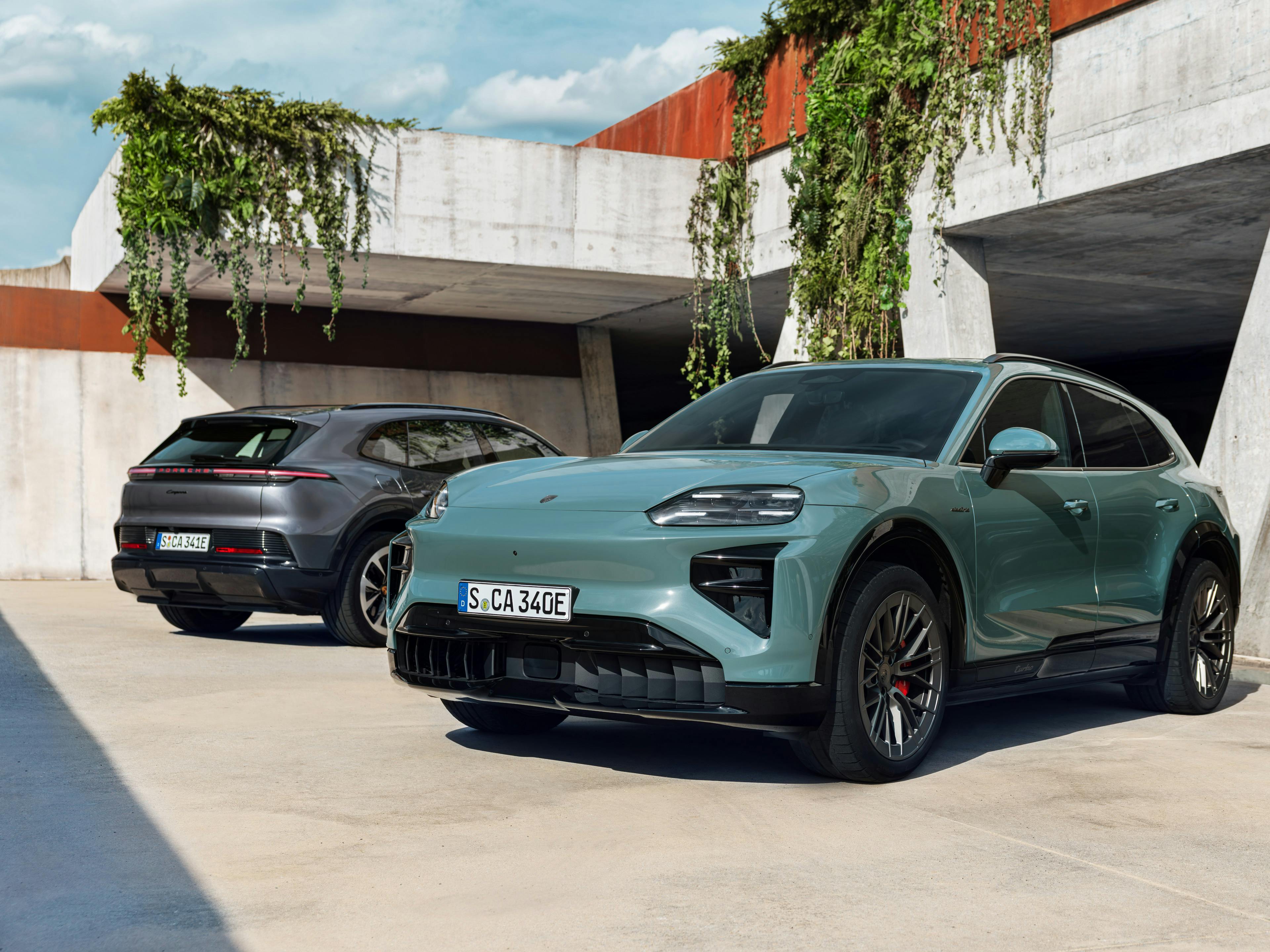 Two Porsche electric SUVs parked outside a modern concrete building with hanging greenery; a light green Porsche Cayenne Turbo Electric is in the foreground, and a dark grey Porsche Cayenne Electric is in the background.