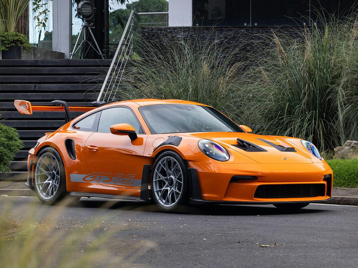 A Porsche 911 GT3 RS in Paint to Sample colour Pastelorange, side and front view, on a street with greenery and modern architecture in the background.