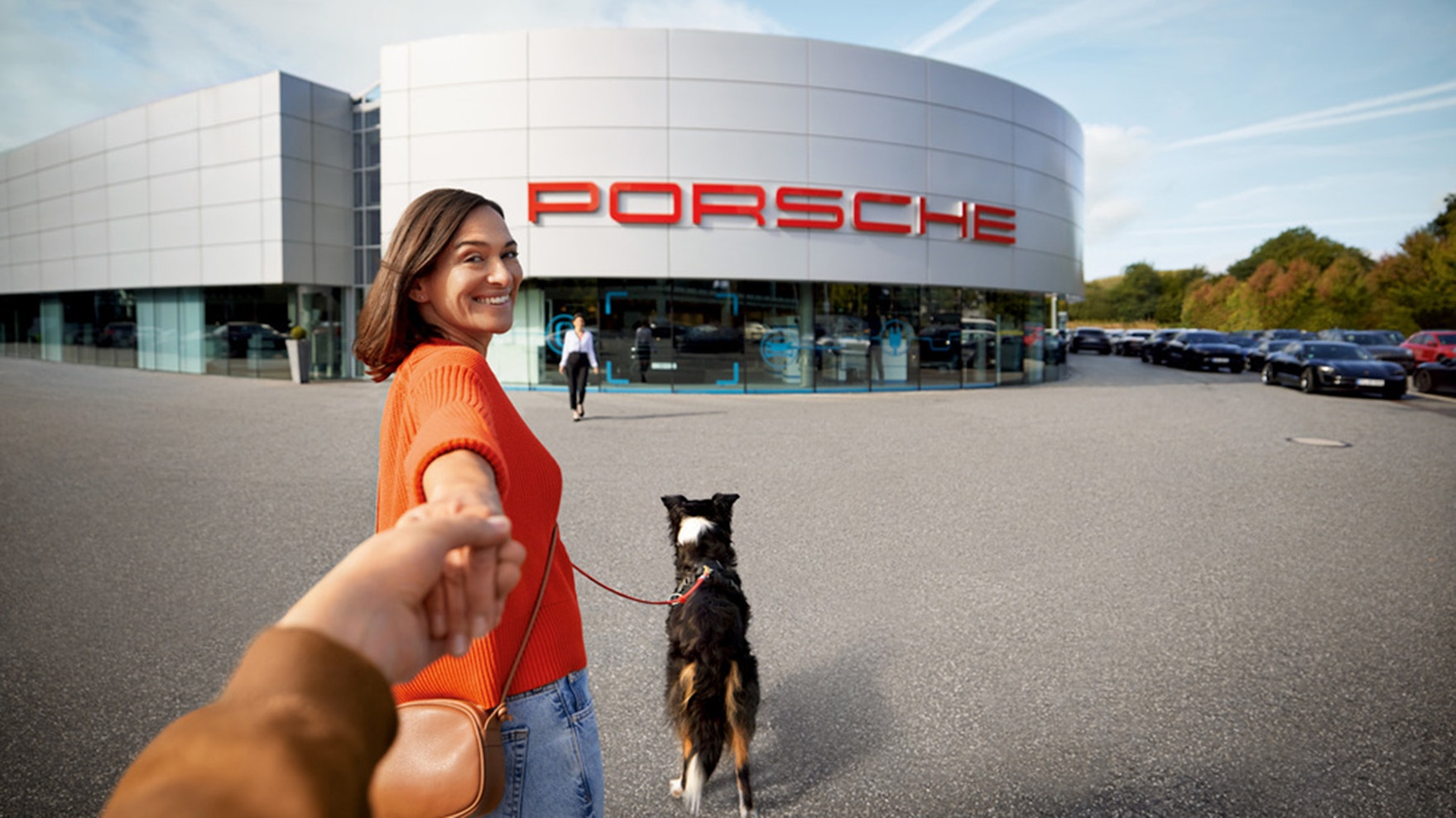 A smiling woman looking back holding hands and carrying a dog in a leash in front of a Porsche Center.