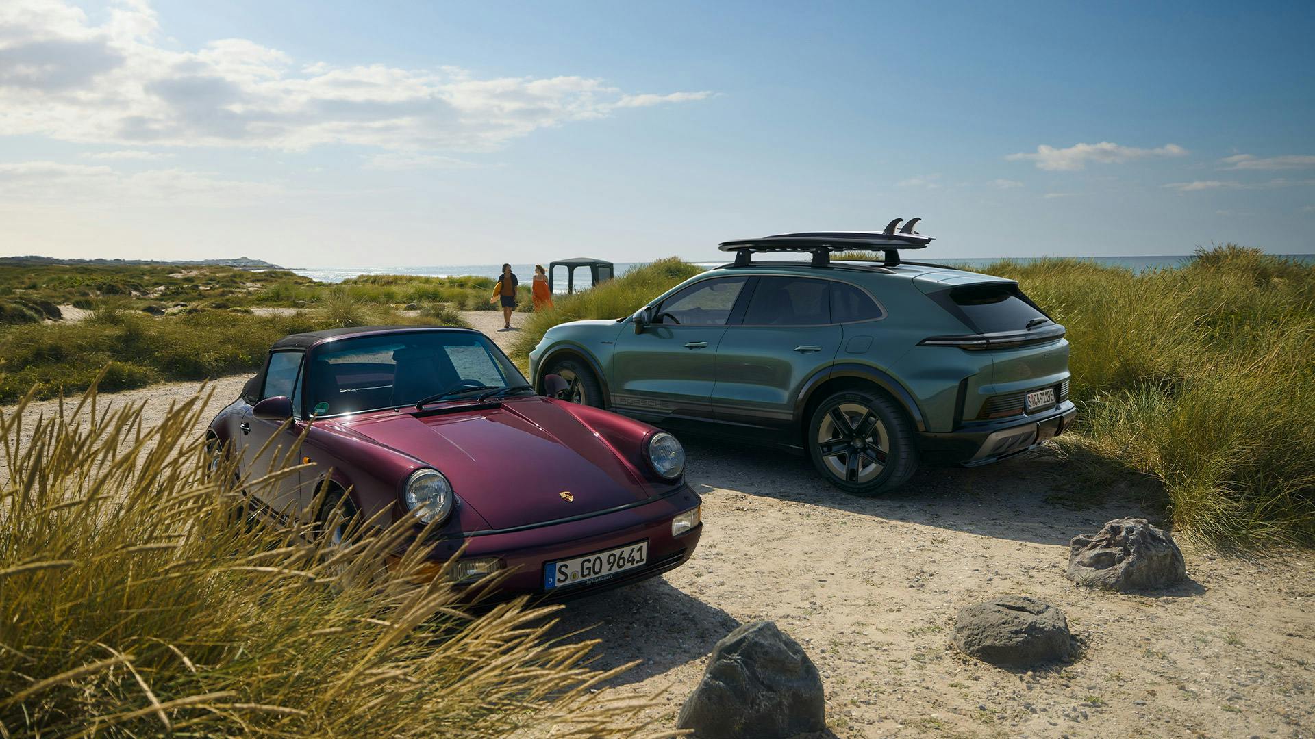 A Cayenne Turbo E-Hybrid with roof cage and surfboard stands alongside a classic 964 Cabrio in the dunes. A Porsche pavilion can be seen in the background.