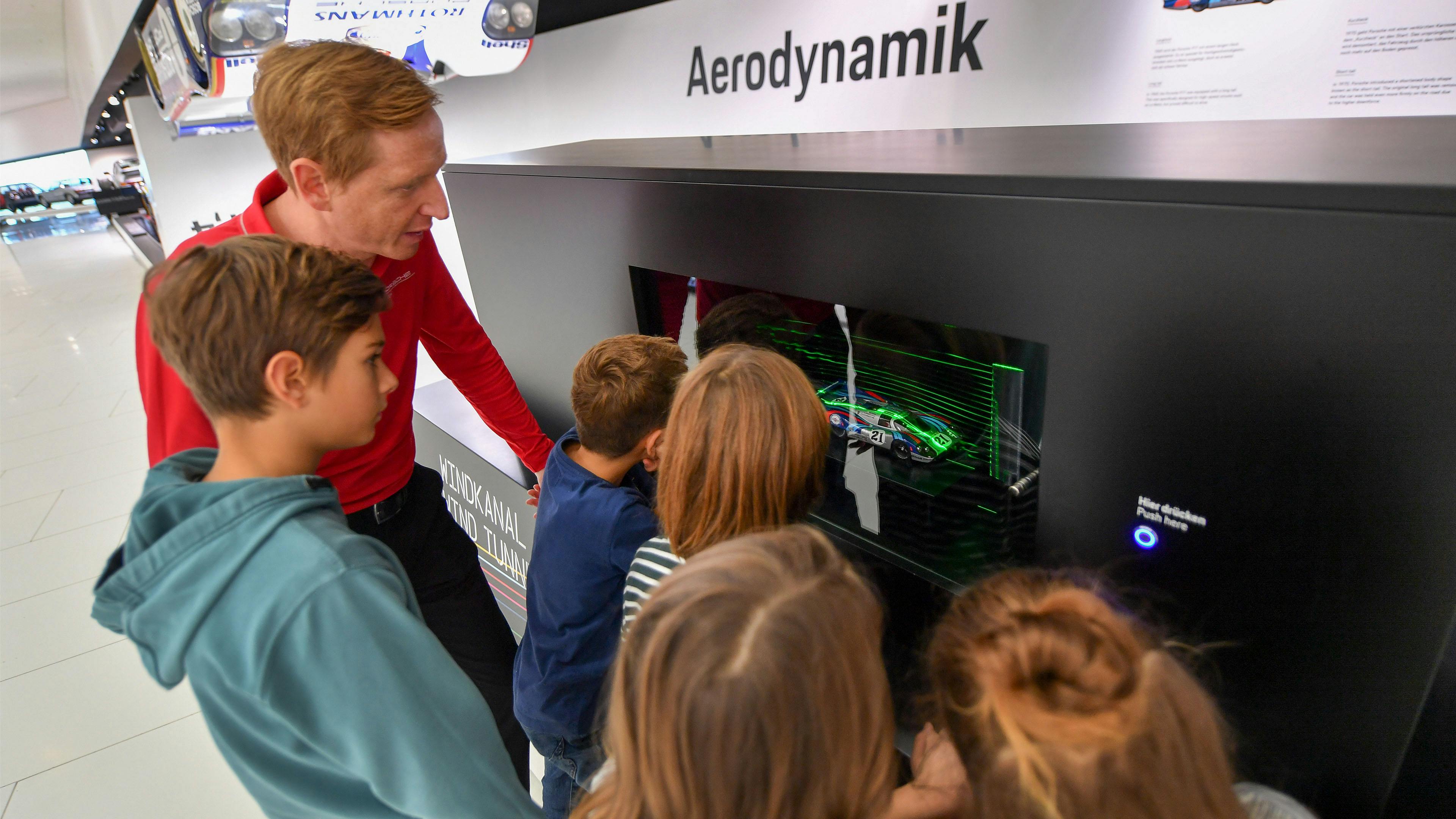 Eine Gruppe von Kindern und ein Porsche Museums Mitarbeiter vor einem Modell zur Darstellung der Aerodynamik