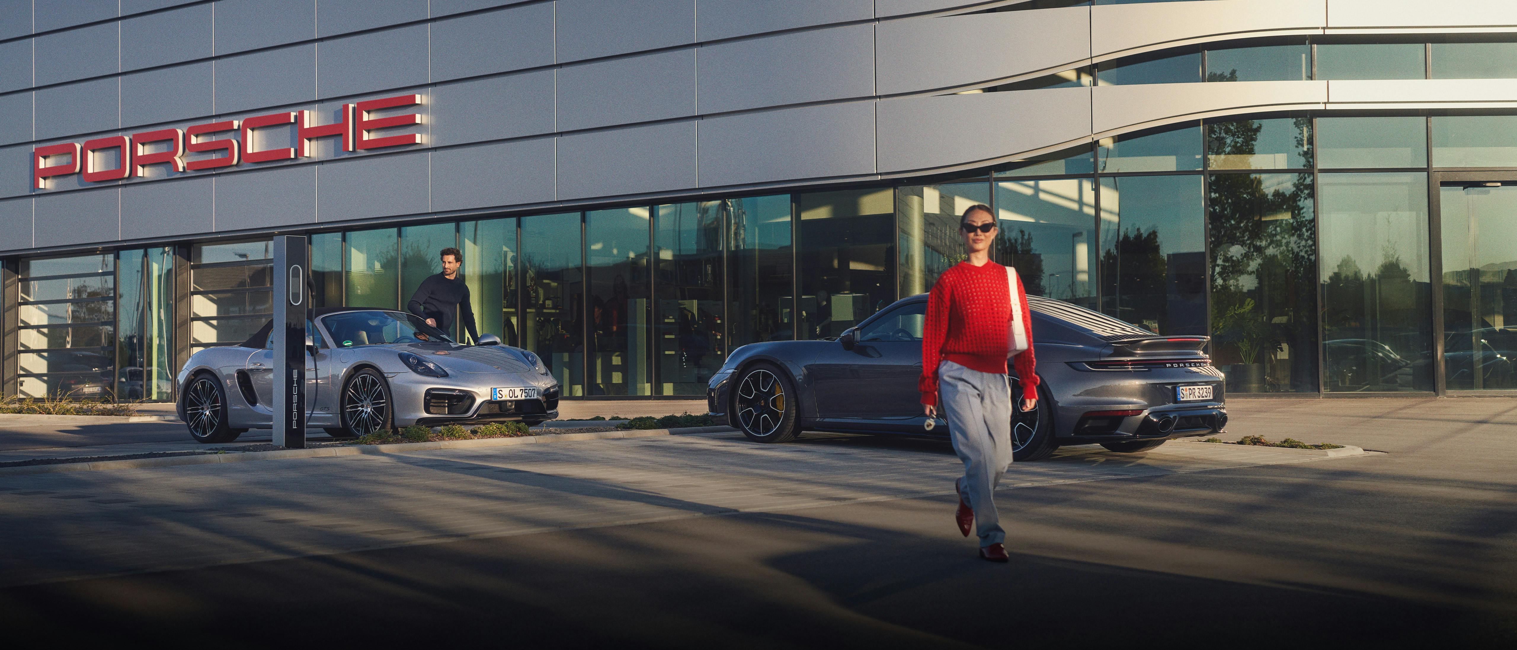 Uma mulher atravessando a rua e dois Porsches estacionados em frente a uma concessionaria Porsche.