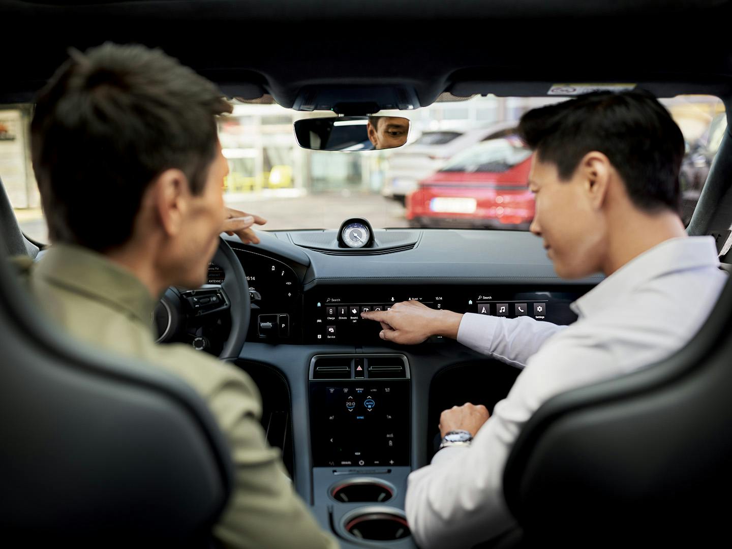 Two men are shown in the interior of a Porsche. The man in the passenger seat points his finger at the center console of the car.