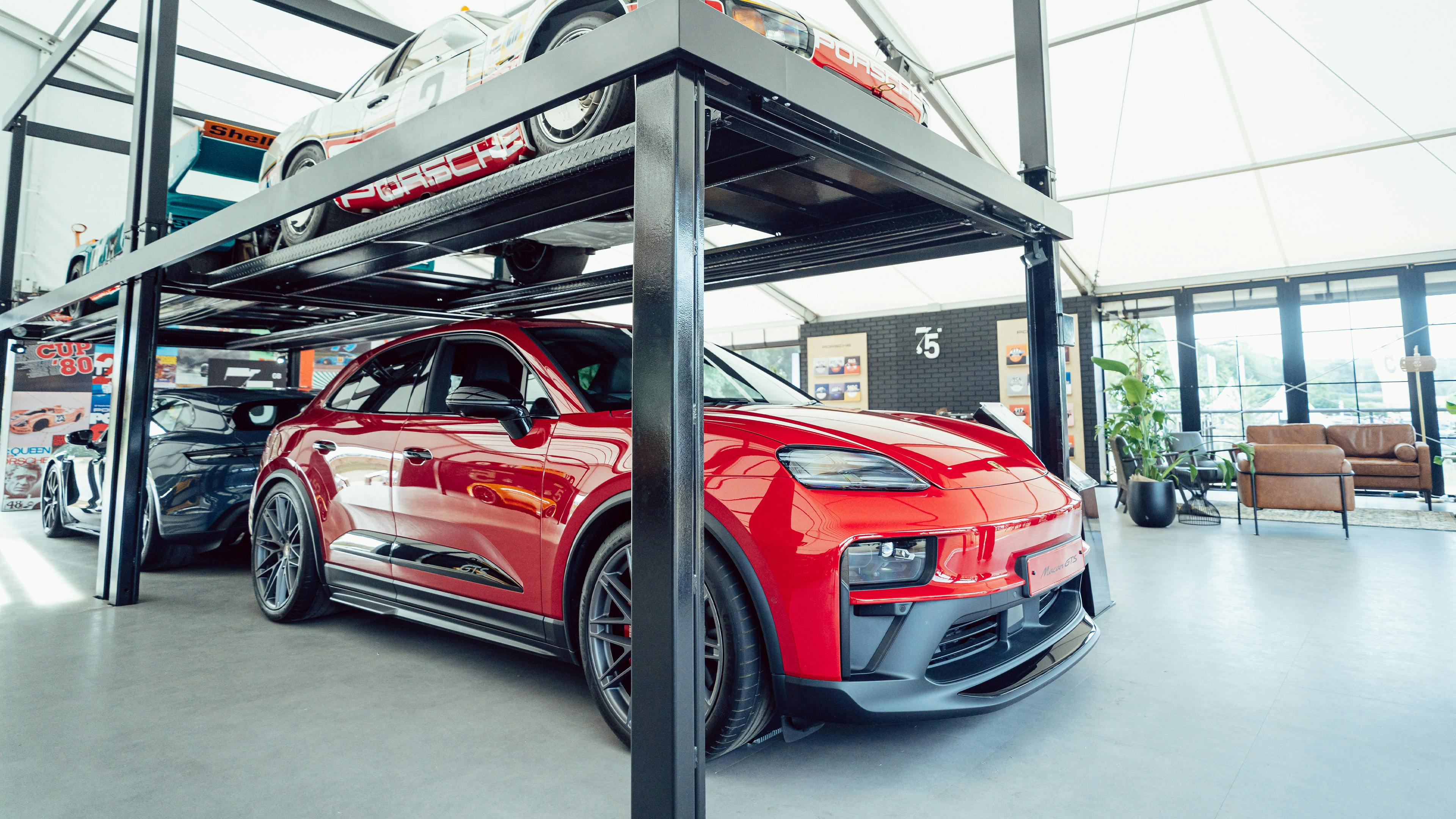 Indoor Porsche display with a race car raised on a car lift above a red Cayenne, showcased within a glass-walled pavilion.