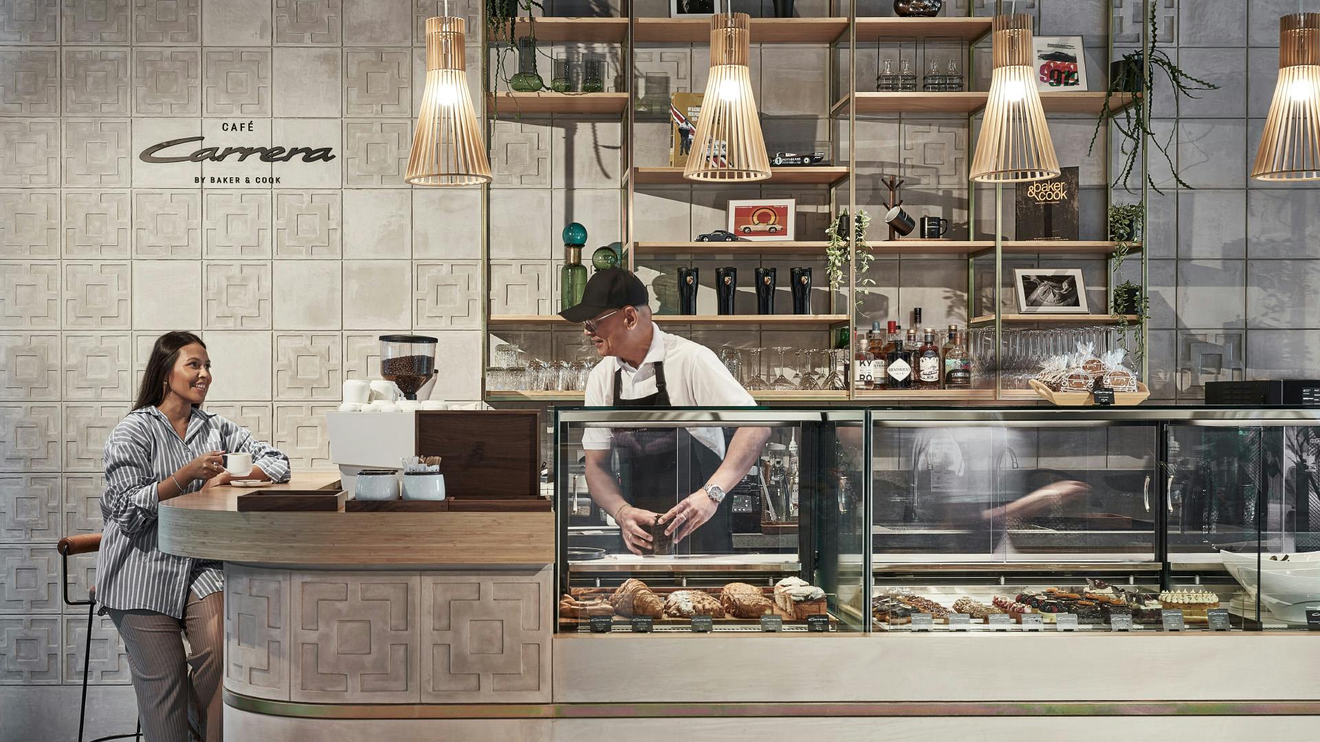 A woman ordering coffee at a stylish café counter labeled “Carrera”, with a barista preparing pastries behind a glass display.