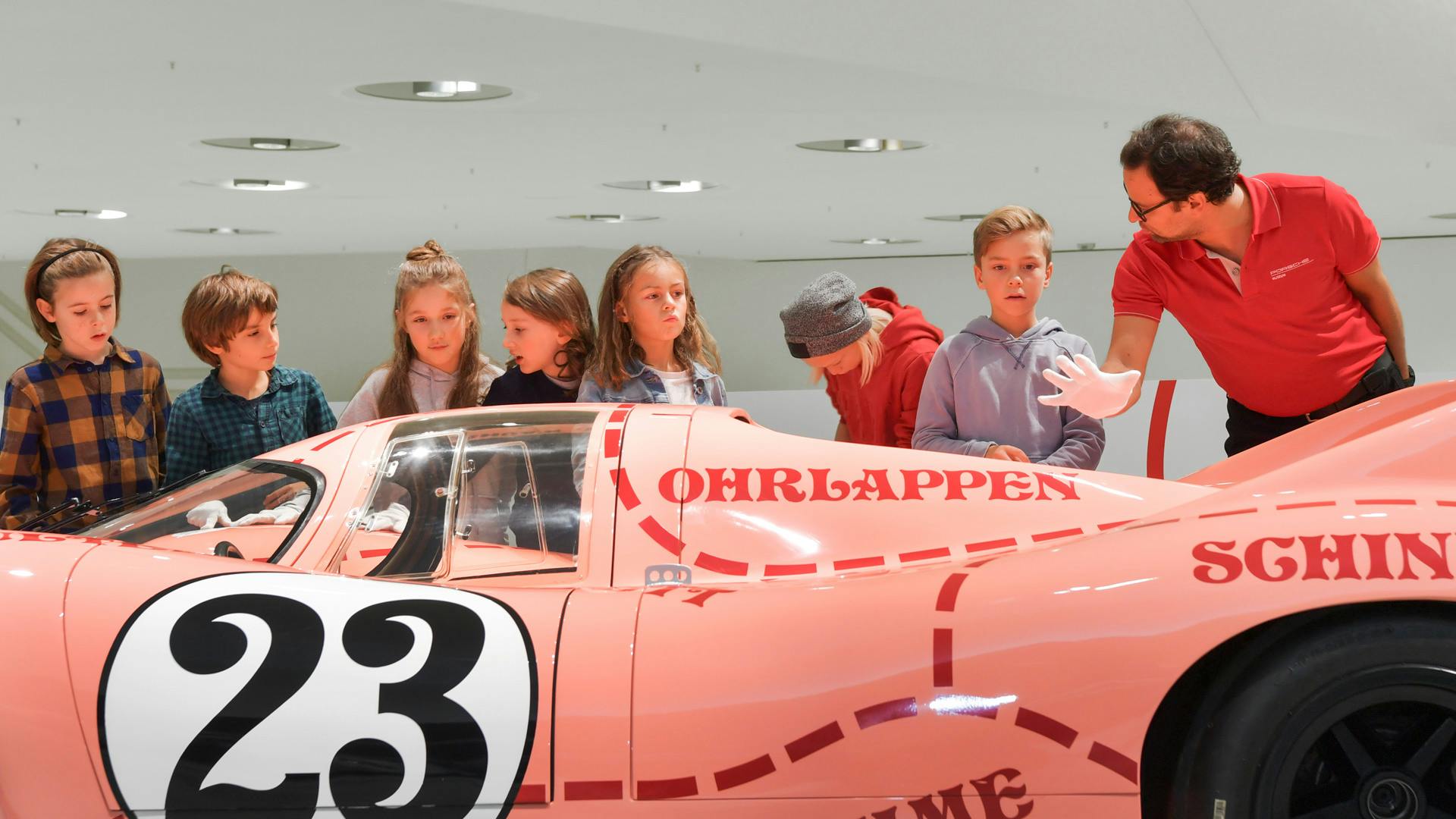 Eine Gruppe von Kindern vor einem rosanen Porsche-Exponat mit einem Museumsführer, der über das Auto spricht