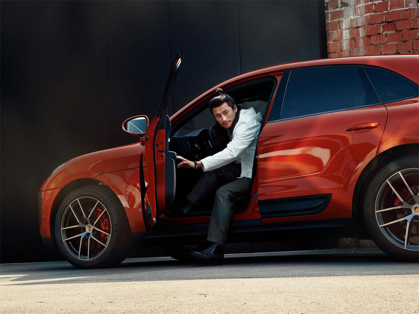 An asian man stepping out of an orange Porsche Macan in the city.
