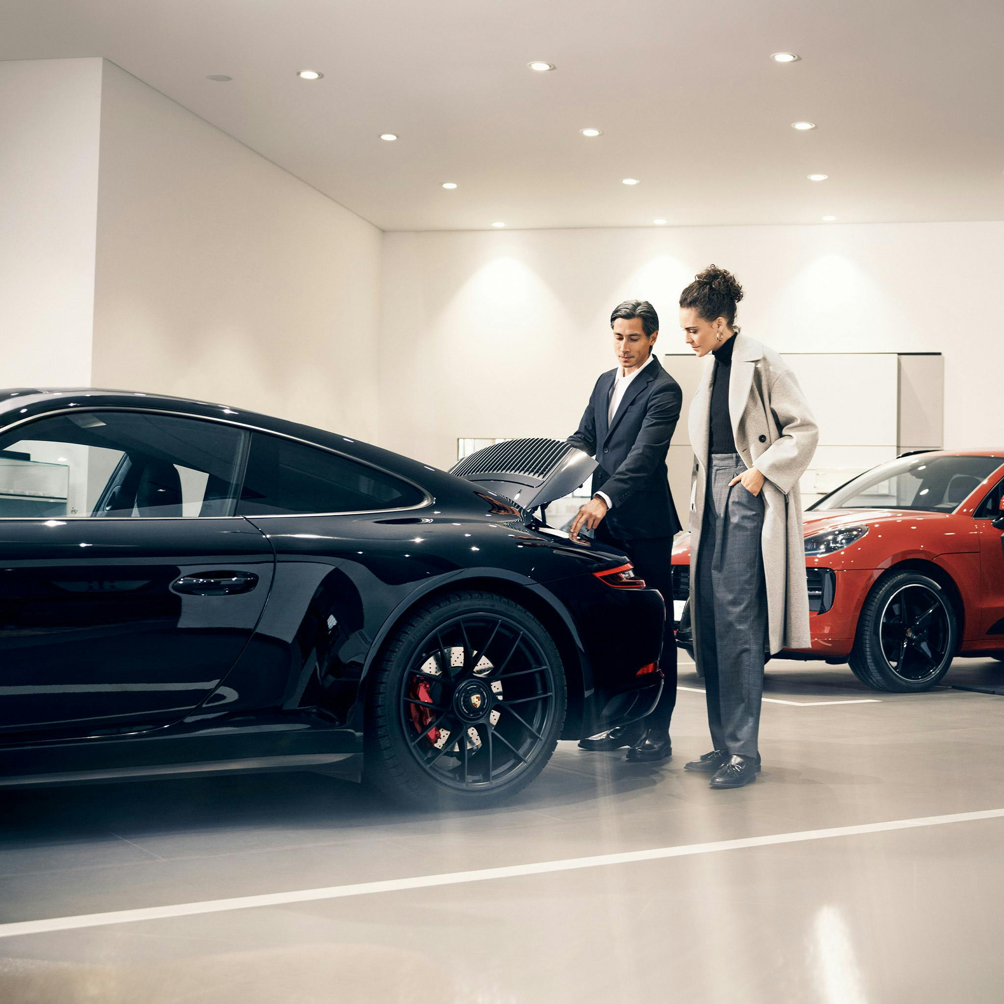 Un homme présente une Porsche noire avec un coffre ouvert à une femme au Centre Porsche. À l'arrière-plan se trouve une Porsche orange.