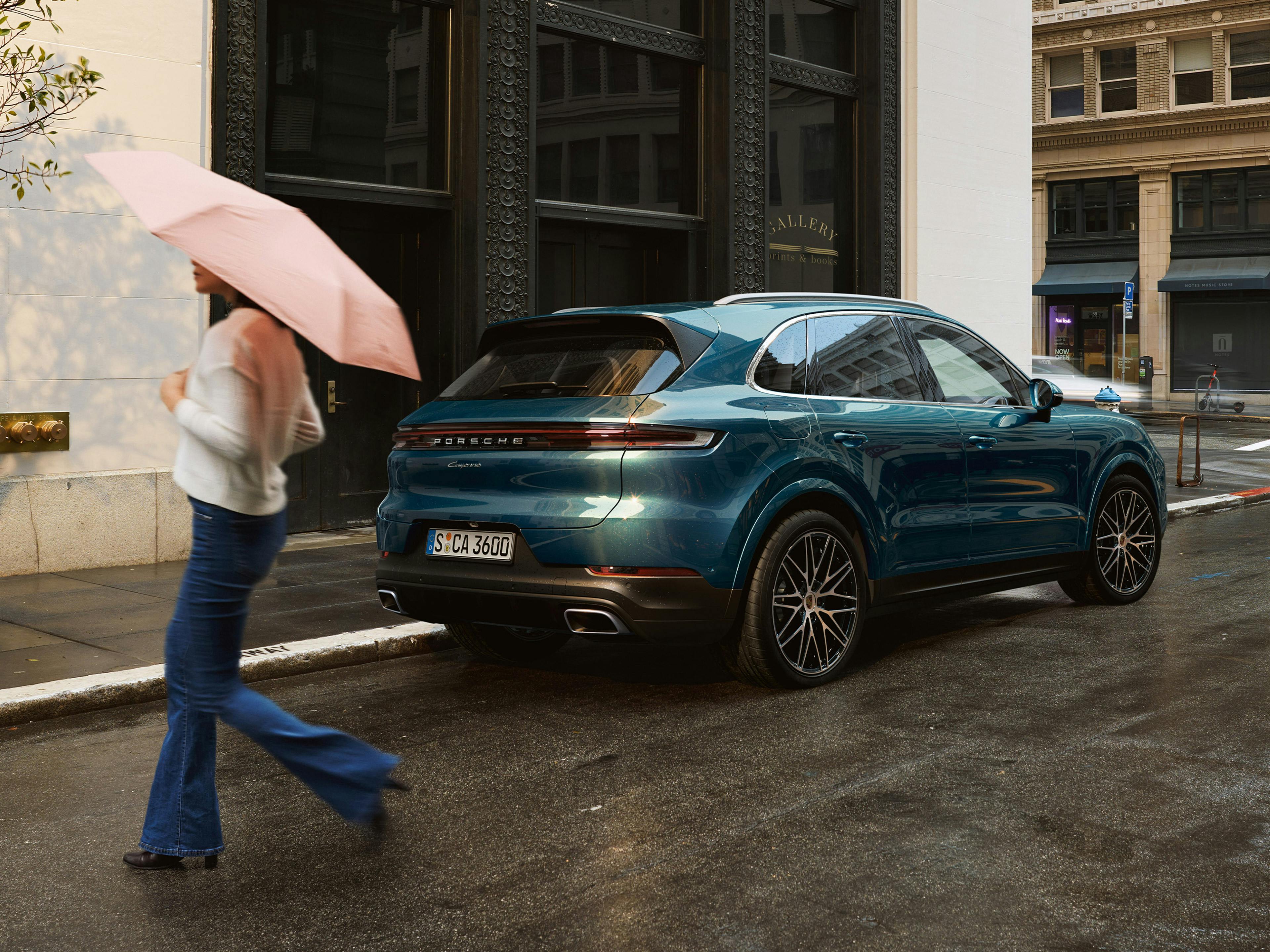 Ein blauer Porsche Cayenne ist an der regennassen Seite geparkt. Eine Frau mit Regenschirm kreuzt die Straße.