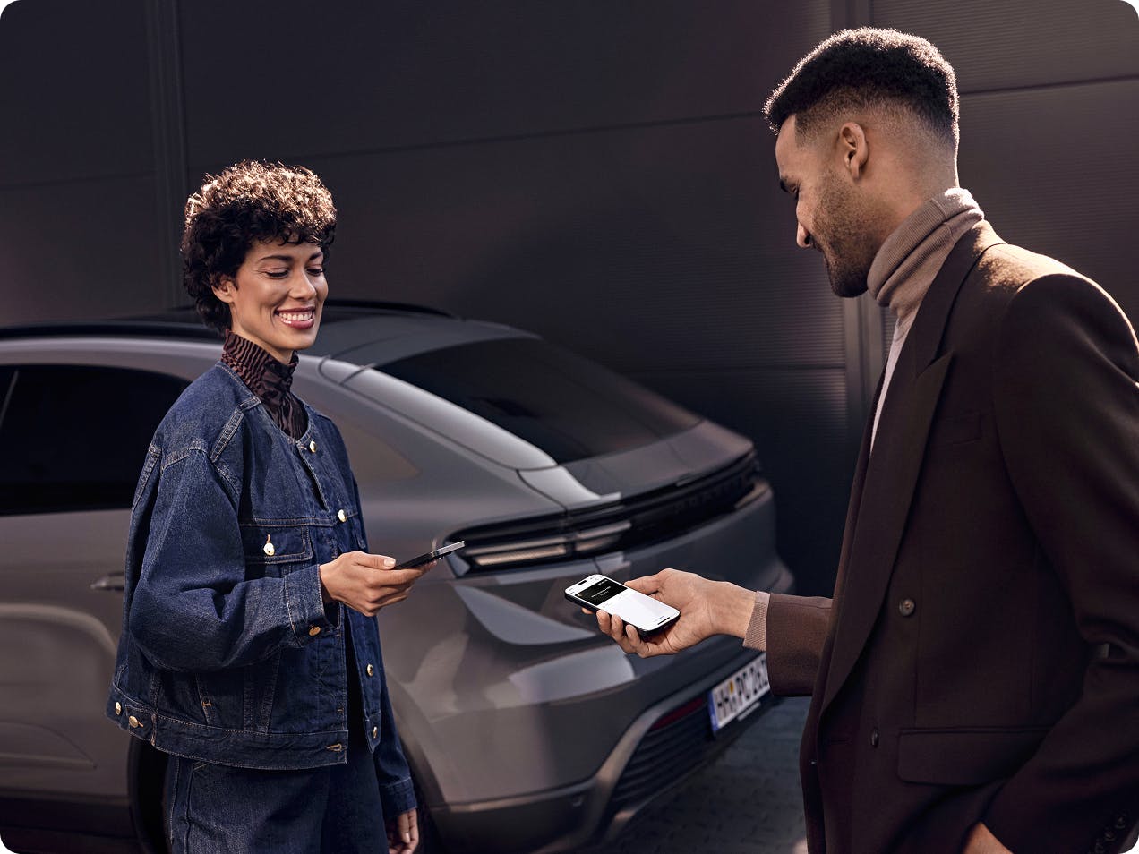 Two people stand facing each other, smiling and looking at their smartphones. A Porsche Macan stands in the background.