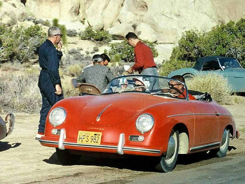 A vintage photograph of a red Porsche 356 convertible with passengers, surrounded by people in a rocky landscape.