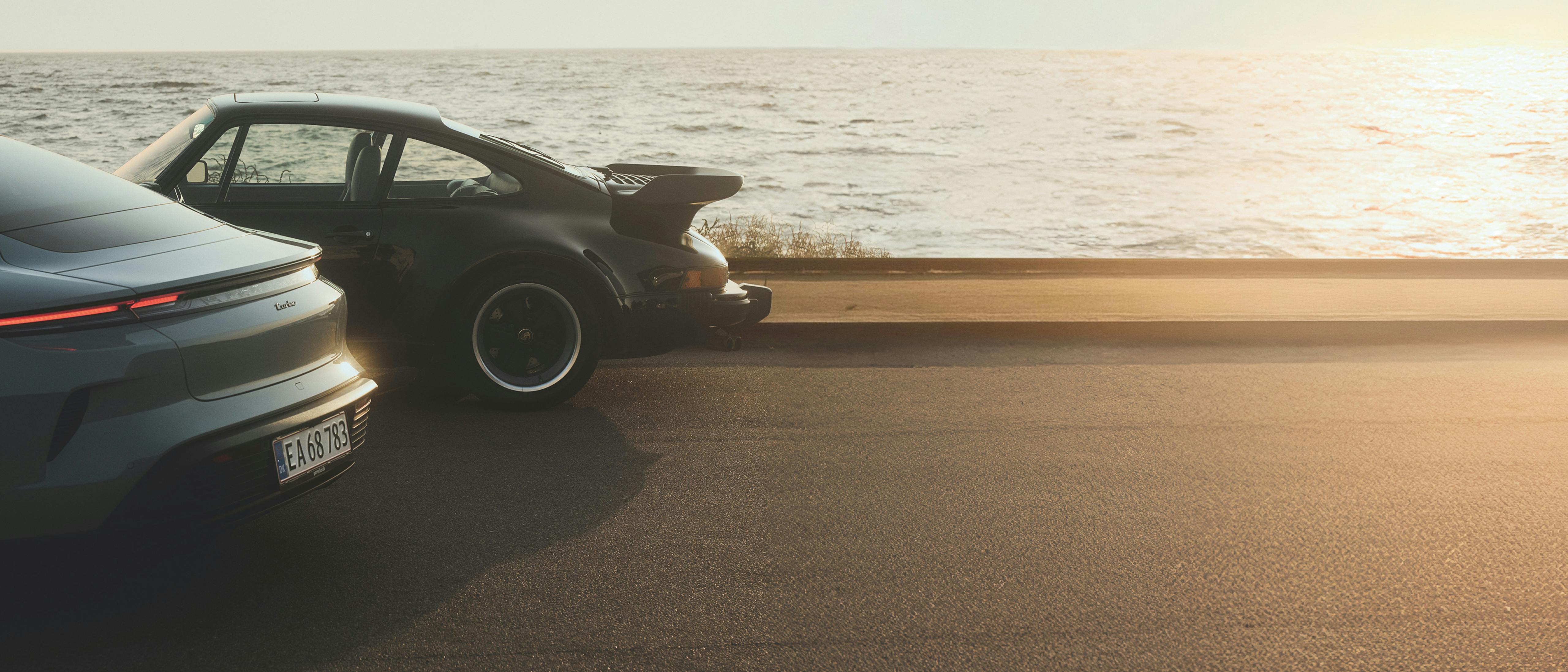 A modern Porsche and a classic 911 Turbo with its distinctive whale tail spoiler, parked together on a coastal road at sunset with the sea behind them.