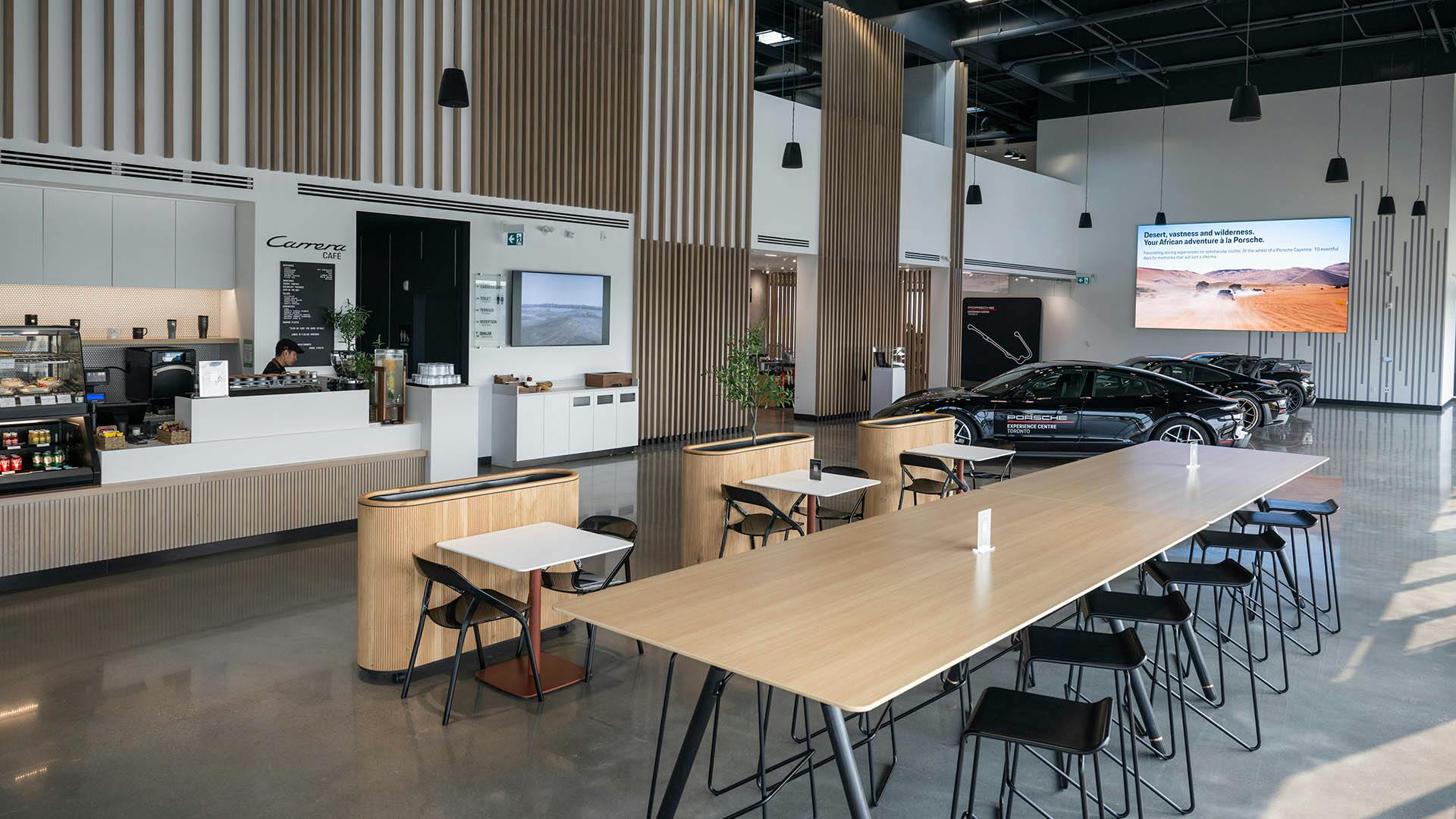 A large modern interior space with a café, large wooden tables, and Porsche vehicles.