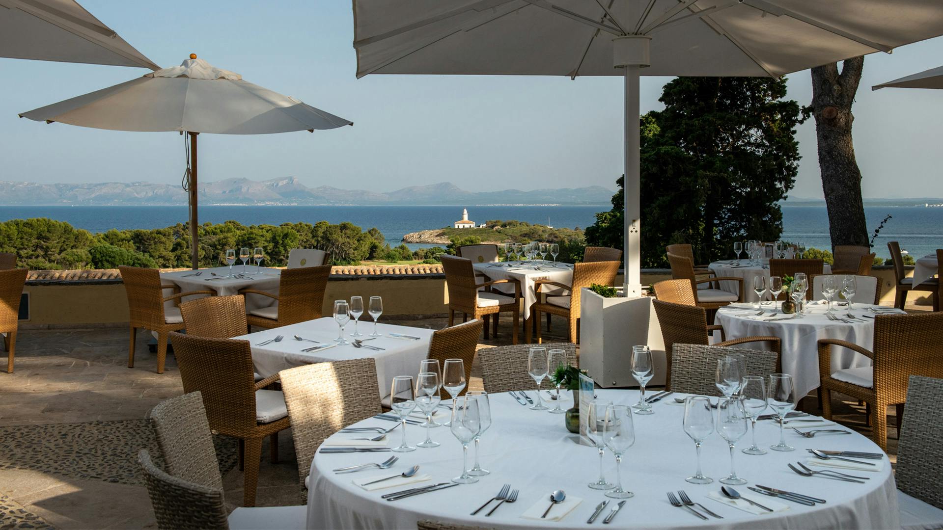 Gedeckte Tische auf einer Außenterasse mit Sonnenschirmen und Aussicht auf Meer und Bergen im Hintergrund
