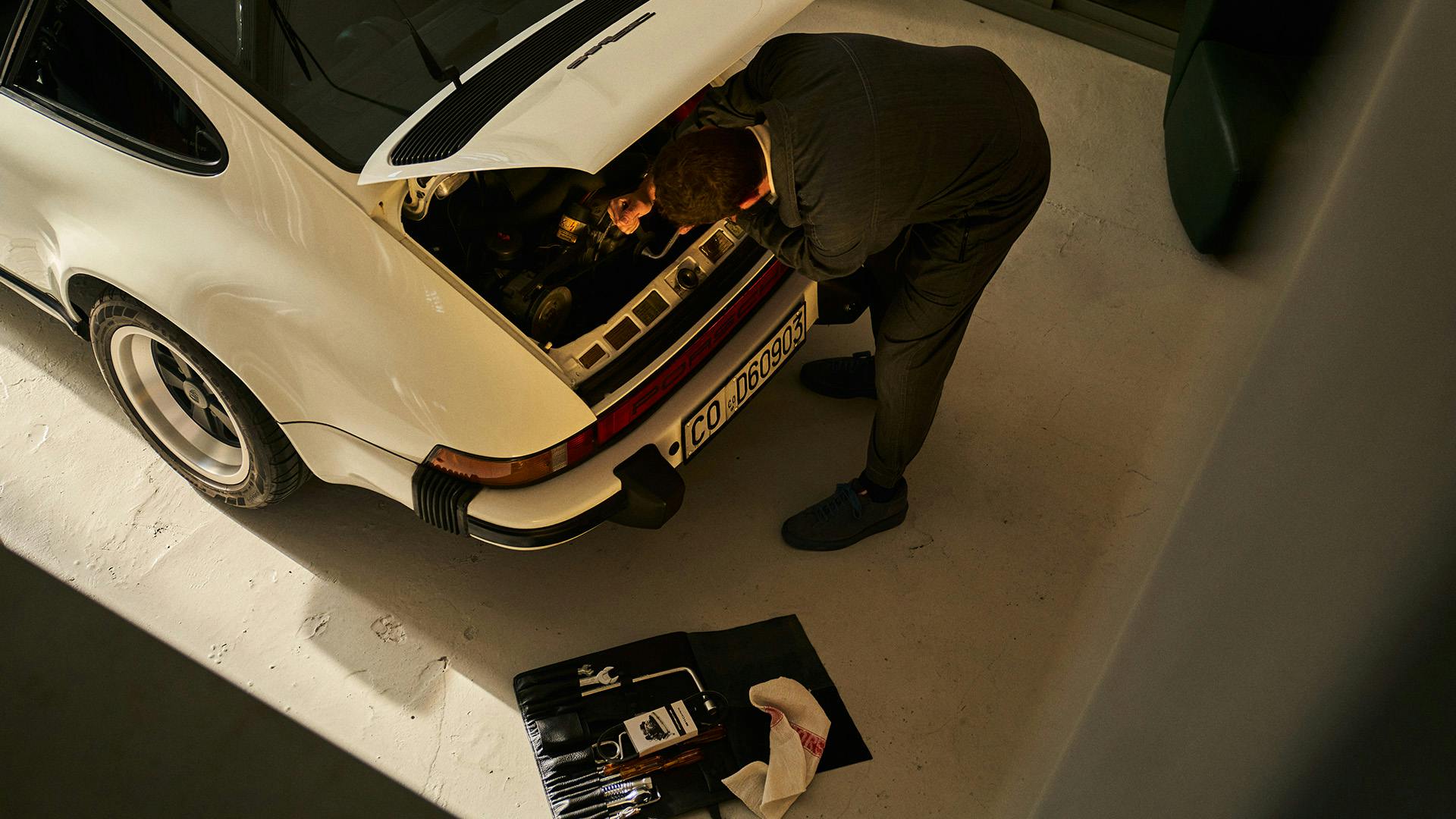 A person is leaning over the open engine compartment of a white Porsche 911 G model; an open tool bag is lying on the ground next to it.
