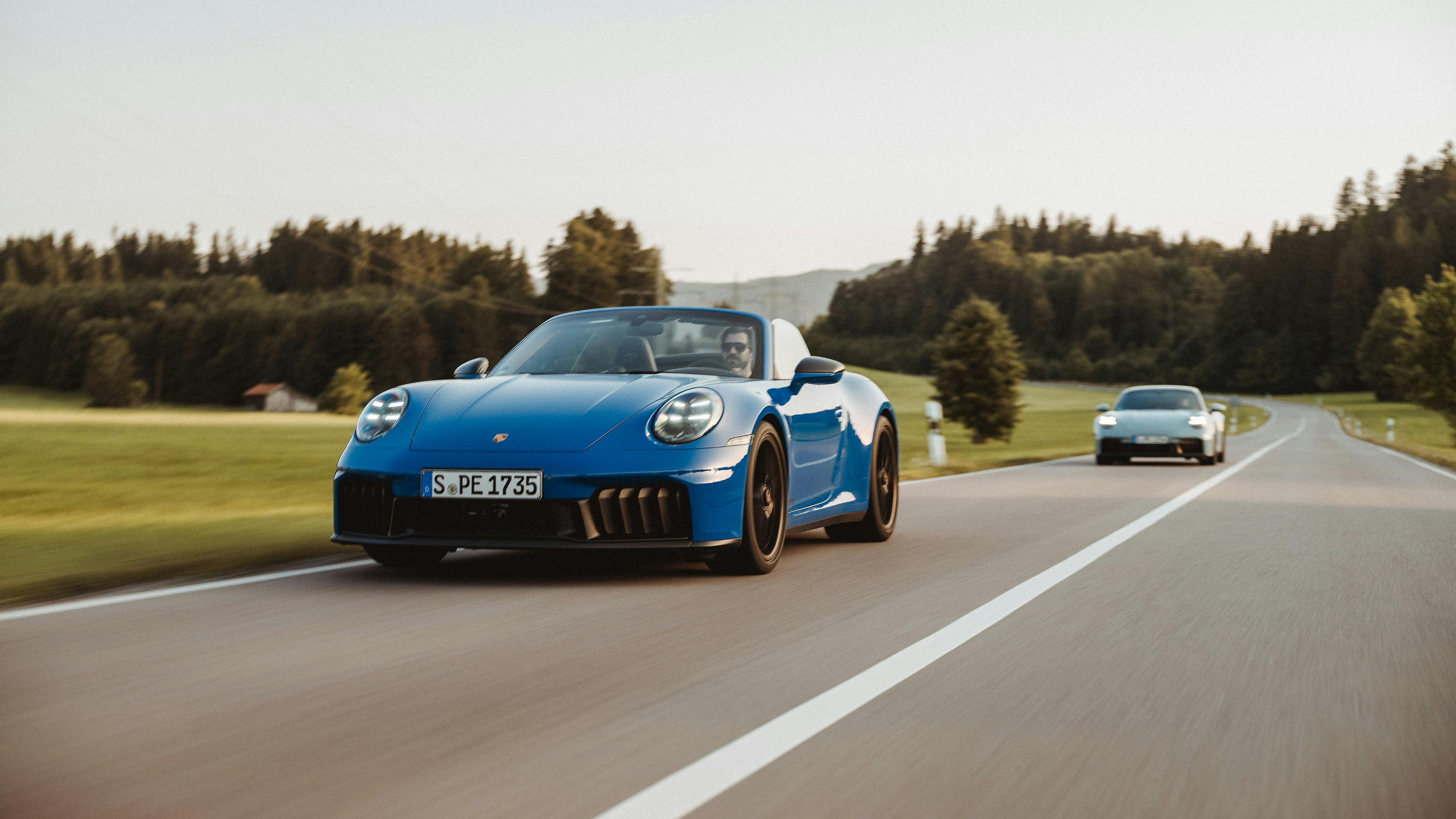 Una Porsche 911 Carrera 4 GTS Cabriolet blu percorre una strada di campagna attraversando un paesaggio verde e collinare, seguita da un altro veicolo.